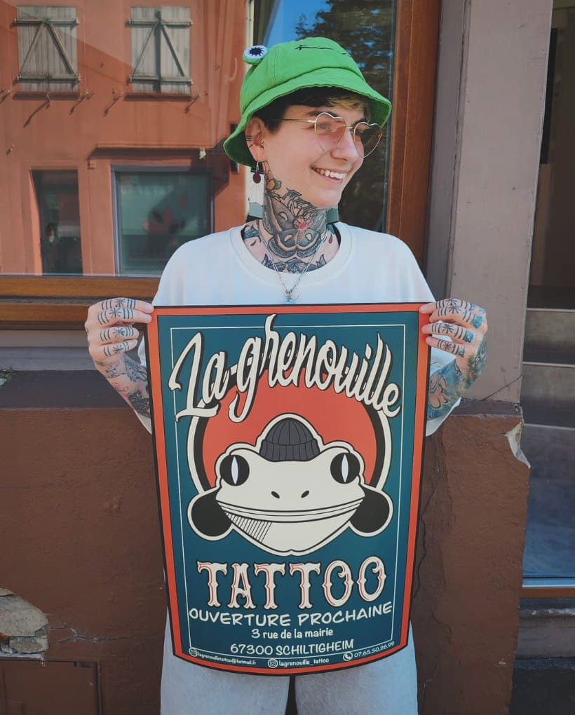 Héloïse Guillement, souriante, bob vert et lunettes cœur, tient l'affiche de La Grenouille Tattoo devant le salon
