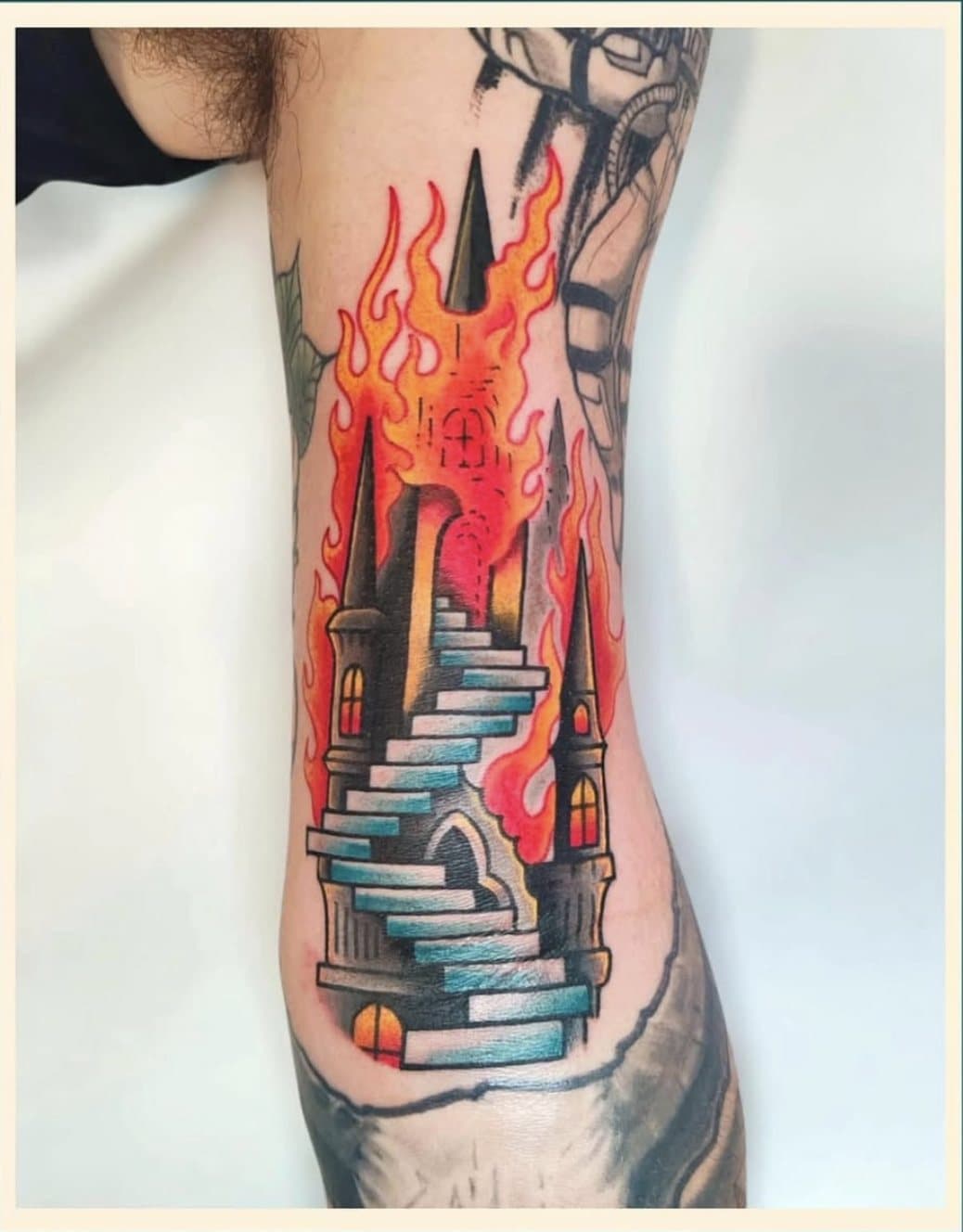 Tatouage couleur sur le bras : un château gothique et son escalier bleu, dévoré par des flammes orange et rouges
