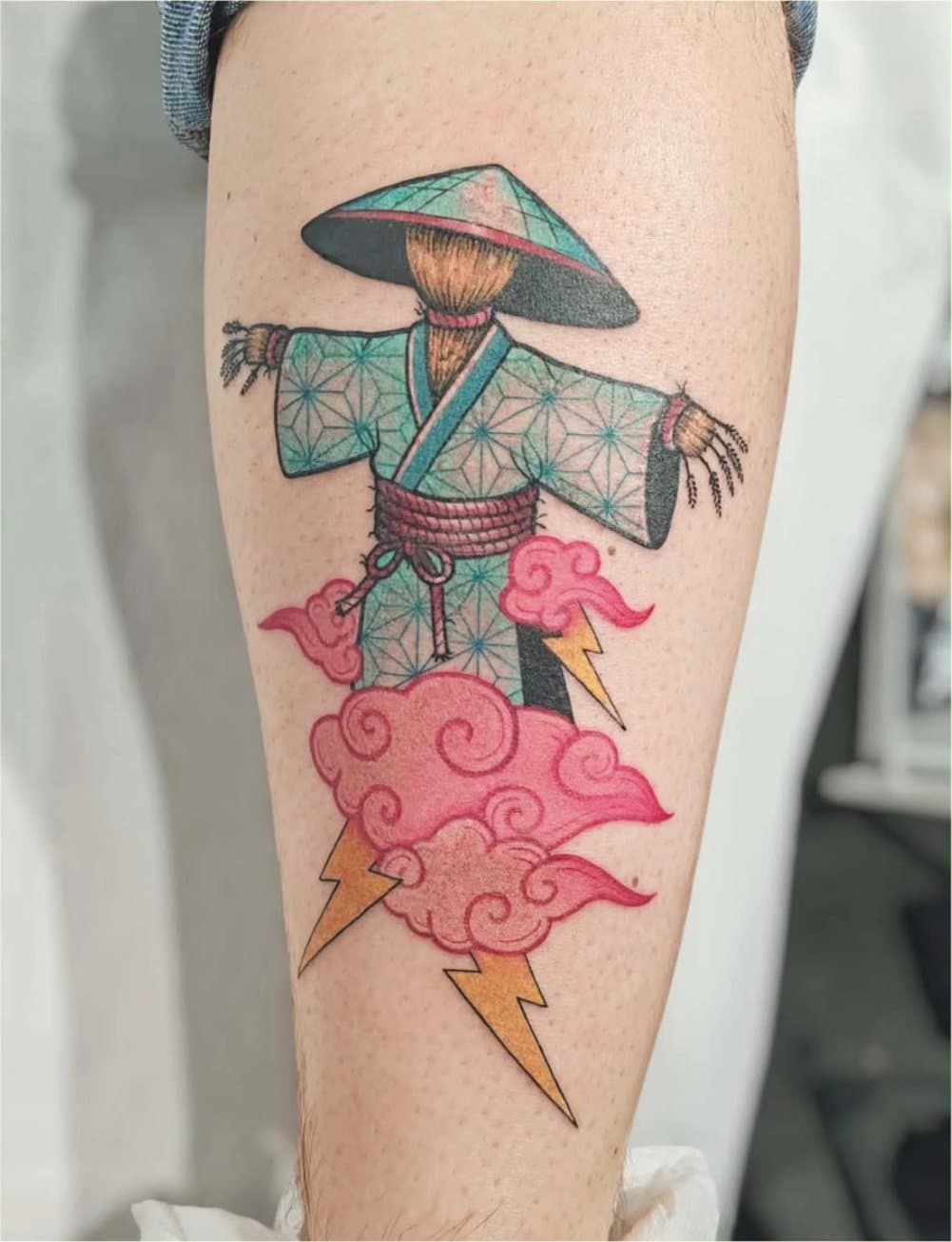 Tatouage couleur : un épouvantail en kimono turquoise à motifs géométriques, chapeau conique, sur des nuages roses et des éclairs jaunes