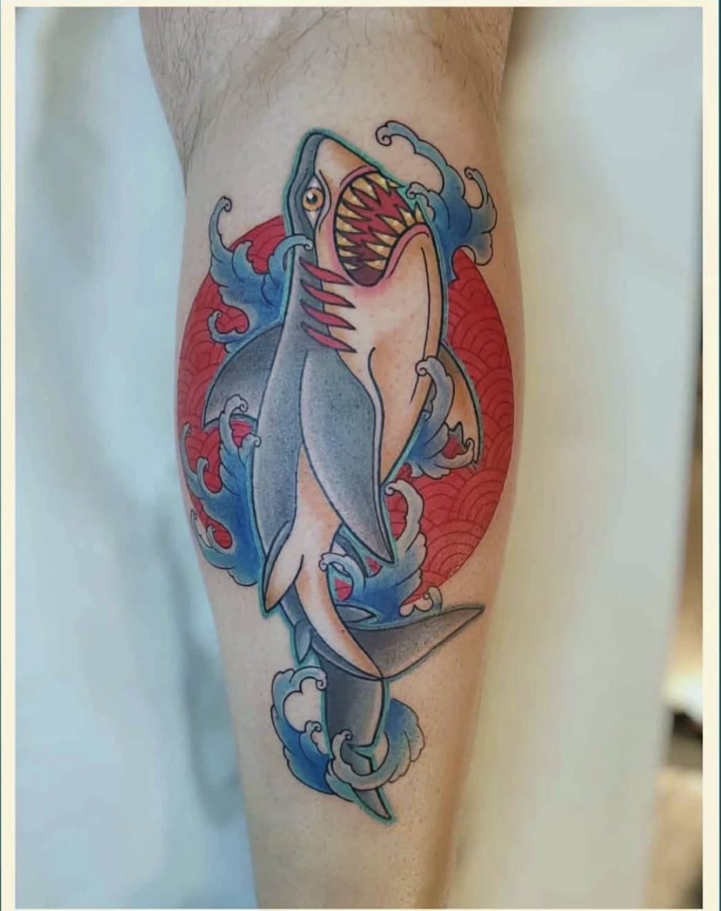 Tatouage couleur sur le mollet : un requin gueule ouverte jaillissant de vagues bleues devant un soleil rouge à motifs