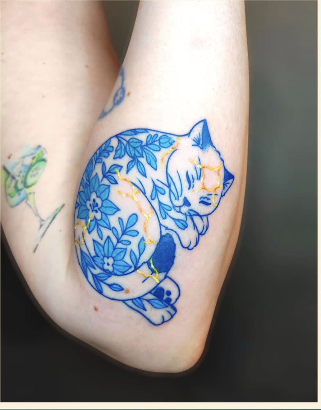 Tatouage bleu sur l'avant-bras : un chat endormi décoré de motifs floraux façon porcelaine, avec des fissures dorées façon kintsugi