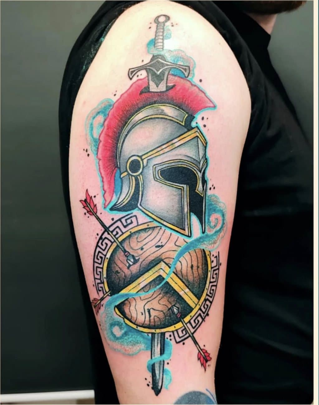 Tatouage couleur sur le bras : un casque spartiate à cimier rouge, épée, bouclier en bois et flèches, avec des volutes turquoise